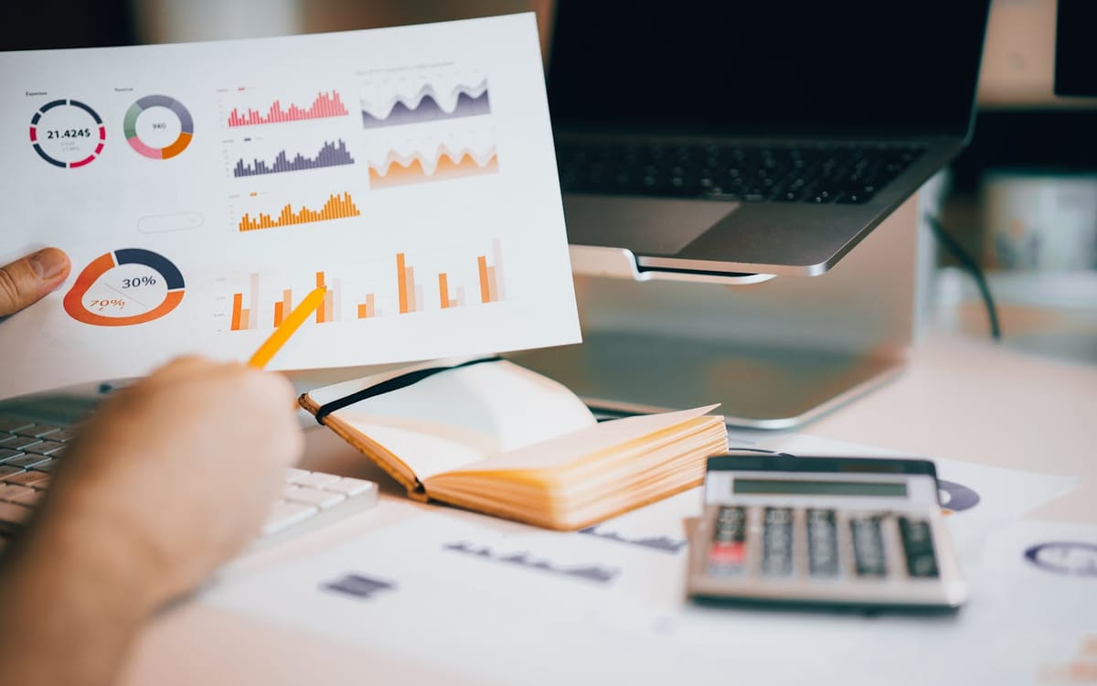 A founder holding a printed dashboard of bar, donut and area charts over a desk with a calculator, pen and open notebook — reviewing the numbers to see which Budget lines change the year's costs