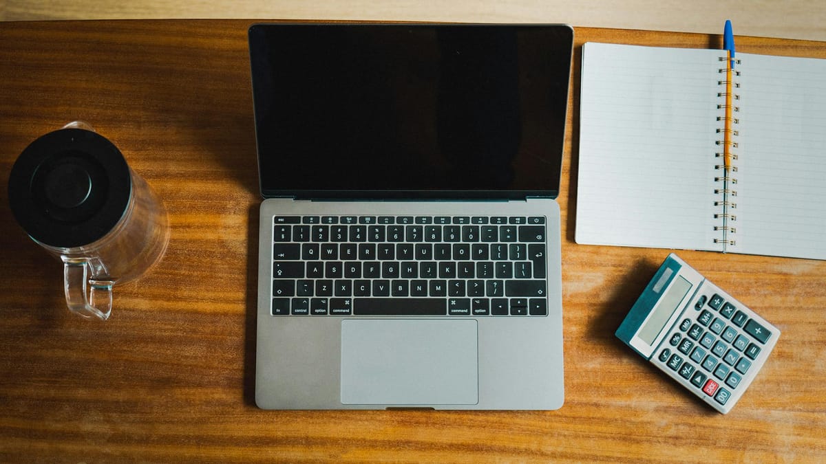 An overhead view of a tidy bookkeeping desk — a laptop, a desk calculator, and an open ledger notebook on a wooden surface — the lean setup a one-founder company runs its books on