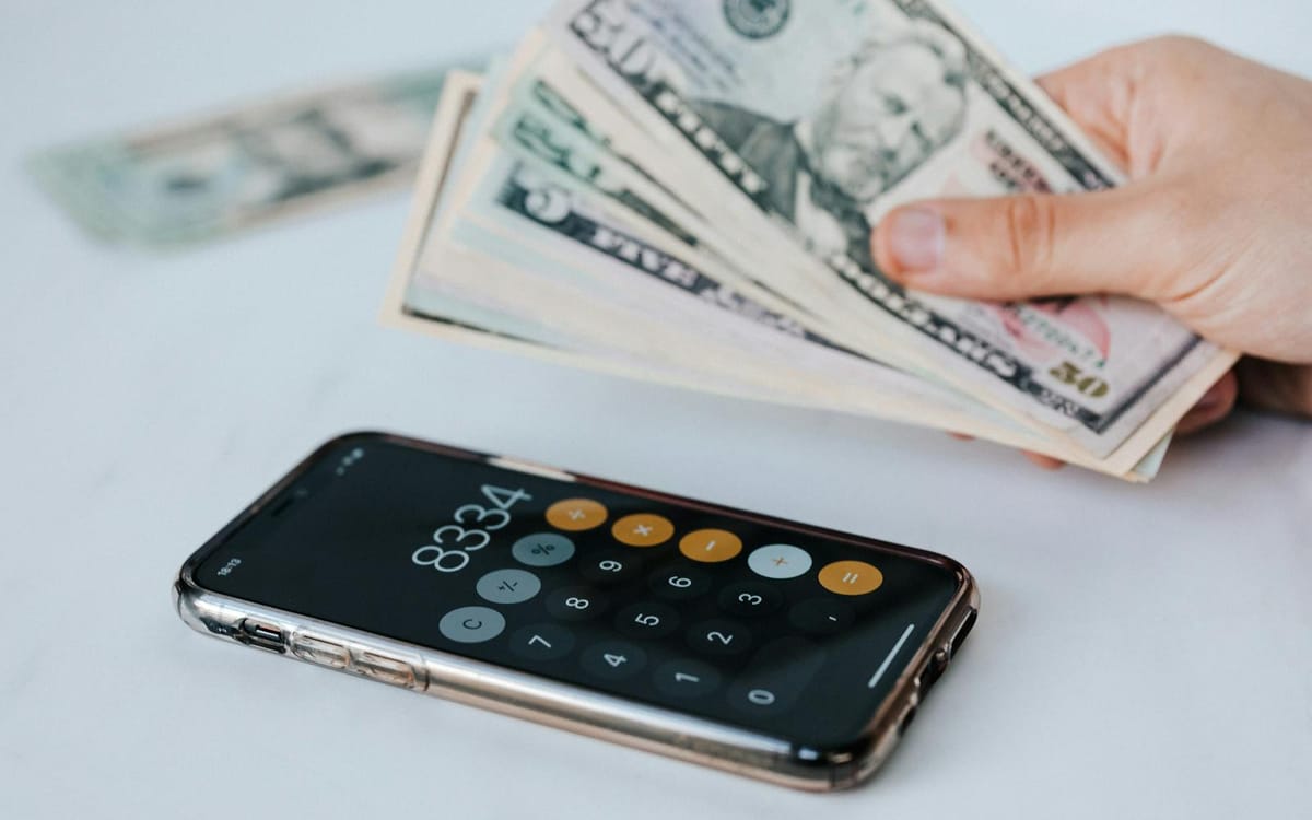 A seller counting US dollar banknotes beside a smartphone showing a calculator — reviewing multi-currency cash flow and supplier costs for a cross-border FBA business