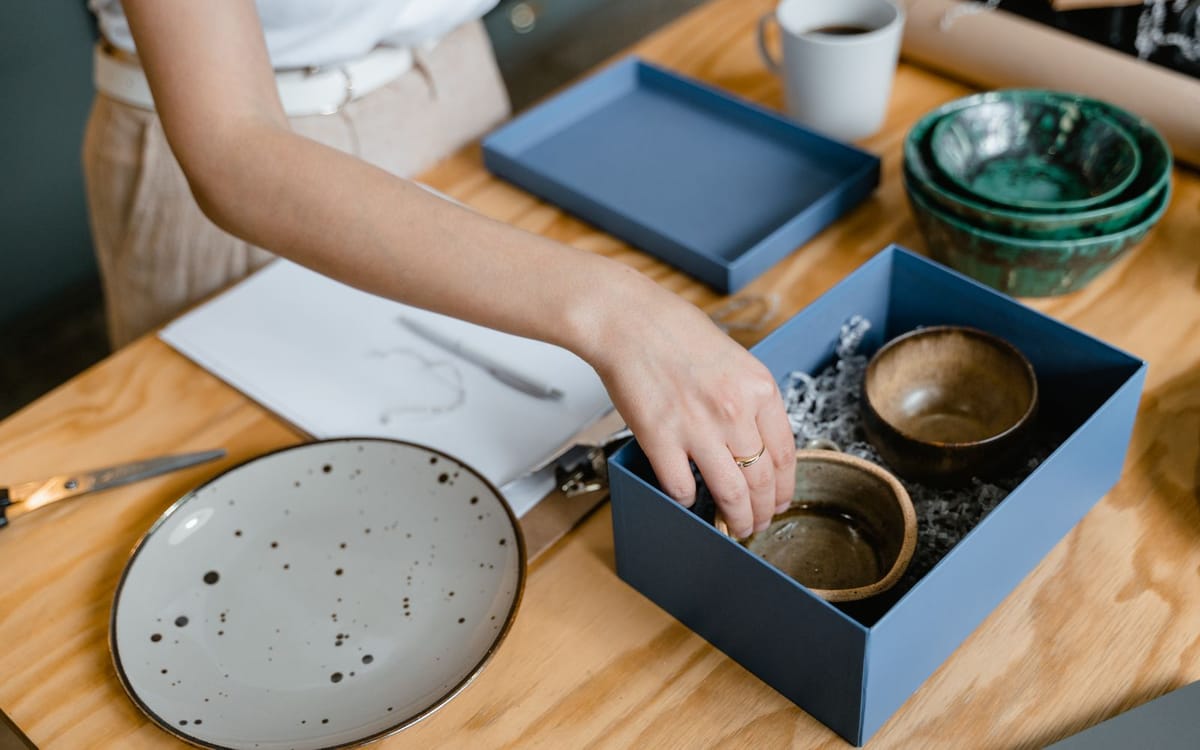 A maker's hands arranging handmade ceramic bowls into a gift box beside a speckled plate and a design sketch — a handmade seller packing products to fulfil and list orders
