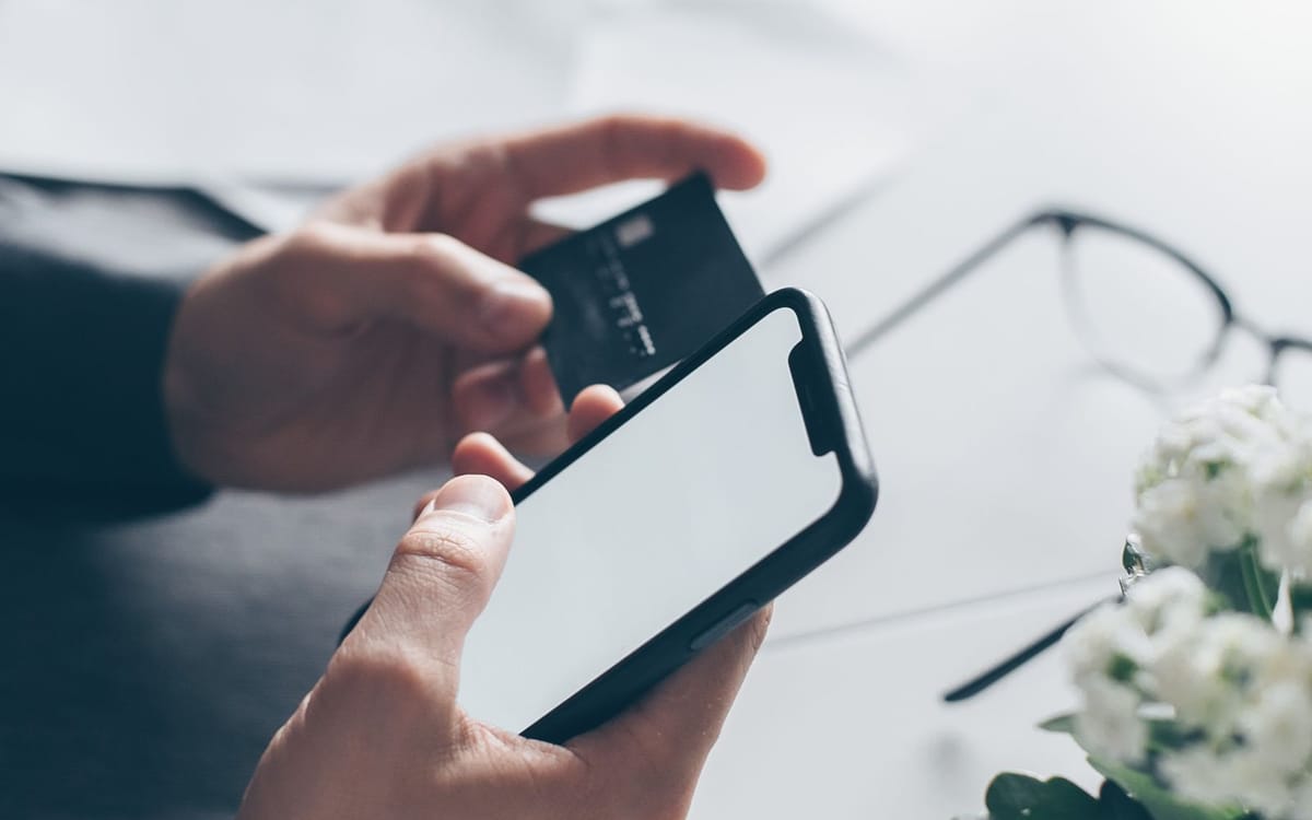 Des mains tenant un smartphone et une carte de paiement au-dessus d'un bureau lumineux — banque multi-devises et facturation internationale gérées à distance via une société à Hong Kong