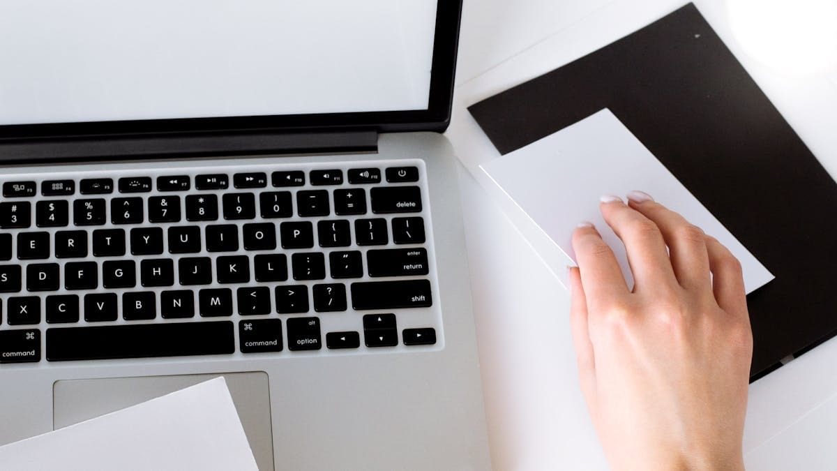 Vue en plongée d'un bureau blanc épuré et lumineux avec un ordinateur portable ouvert et une main sur le pavé tactile — un espace de travail propre et indépendant du lieu, pour un expatrié qui gère son activité à distance