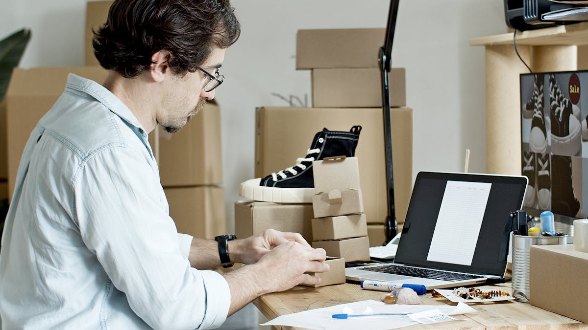 An online seller packing product boxes at a desk with a laptop and stock around — running cross-border e-commerce fulfilment