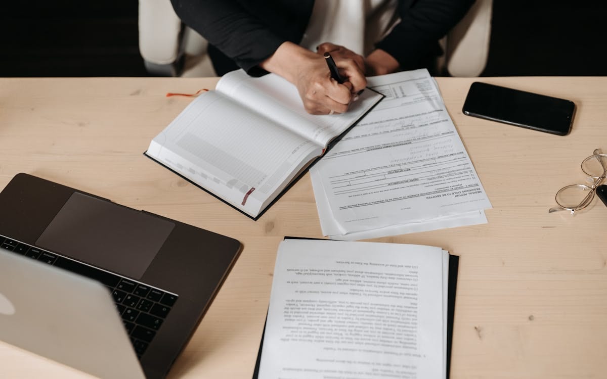 Overhead view of hands signing company documents beside a laptop, phone and glasses on a wooden desk — reviewing and approving paperwork digitally from abroad